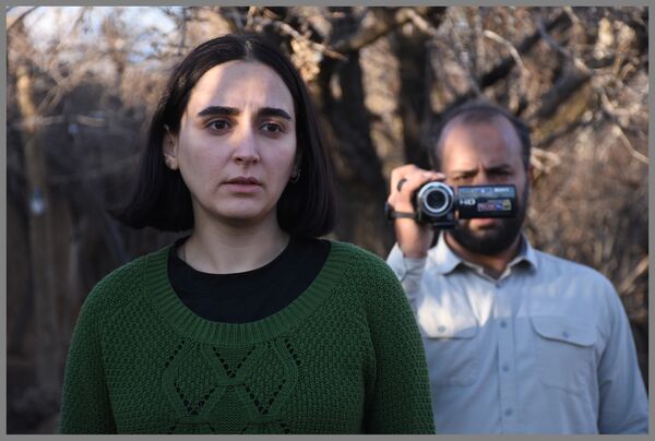 A man with a video camera stands behind a young woman in a woodland. 