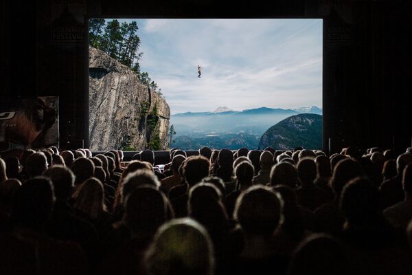 A cinema audience watches a screen. On the screen is a person tightrope walking across an expanse of water. 