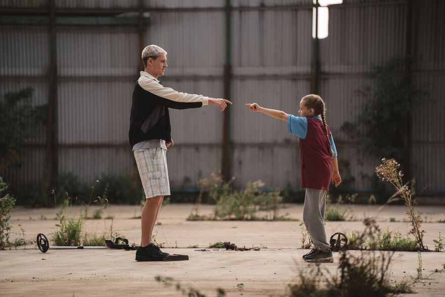 A father and daughter face each other pointing inside an abandoned warehouse. 
