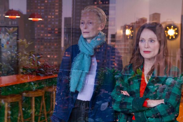 Two women stand together looking out of a window. The city beyond can be seen reflected in the glass. 