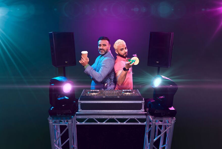 Two men holding up drinks as they stand behind a DJ booth