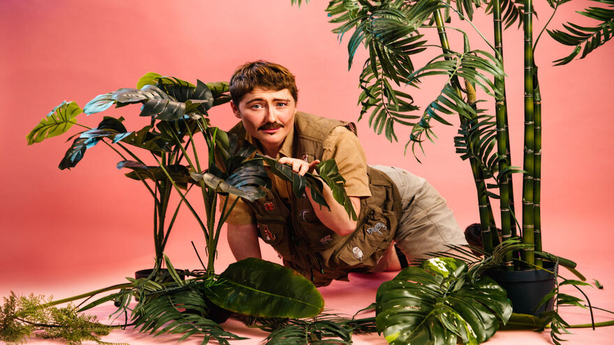 Person wearing safari camouflage outfit and peering from behind faux shrubbery