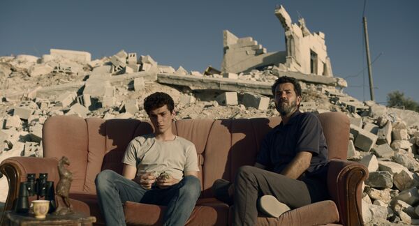 A man and a teenage boy sit on a sofa outside amongst rubble. 