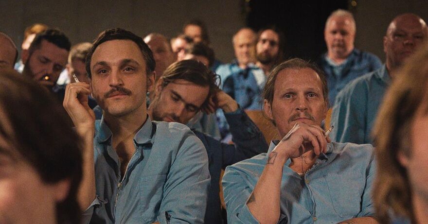 Two men, wearing blue shirts, look forward whilst smoking. They are surrounded by other men sat in rows. 