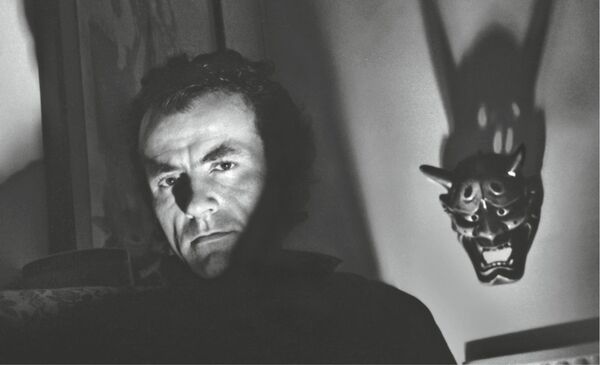 Hugh Cornwell face partially in shadow with an Oni mask in background, Image is in black and white