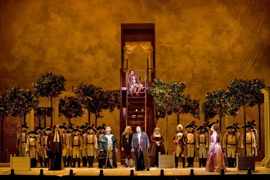 A large ensemble on stage within the setting of an orchard. A man sits on a balcony atop a set of stairs. 